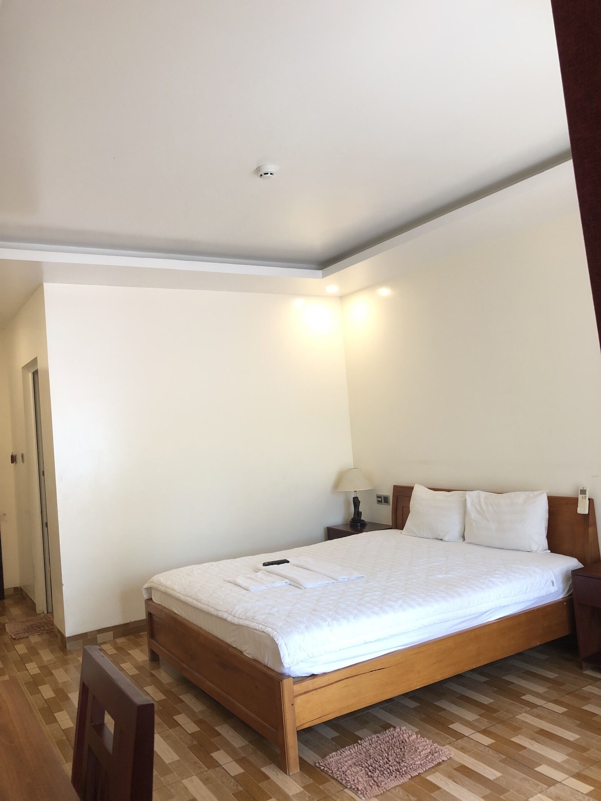 Un dormitorio con cama de madera, ropa de cama blanca, una mesita de noche, una lámpara y una luz en el techo.