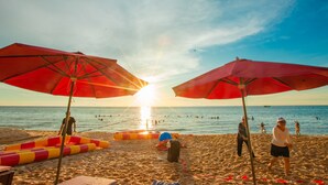 Escena de playa con dos sombrillas rojas grandes, un flotador de piscina y gente nadando y caminando sobre la arena.