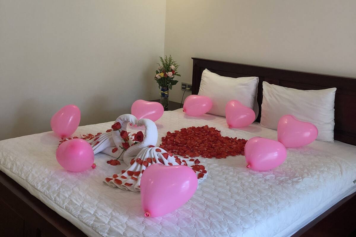 Una cama bien hecha con ropa blanca, globos rosados en forma de corazón y pétalos de rosa dispuestos sobre la cama.