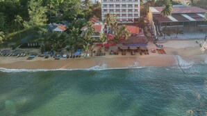 Vista aérea de un hotel costero con varias edificaciones, una playa con tumbonas y palmeras.