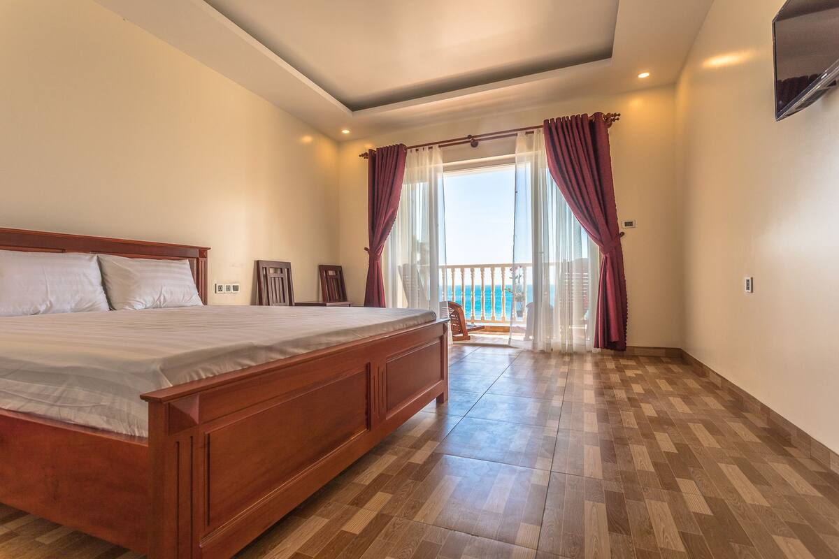Un dormitorio con cama de madera, un balcón con cortinas rojas y vista al mar.