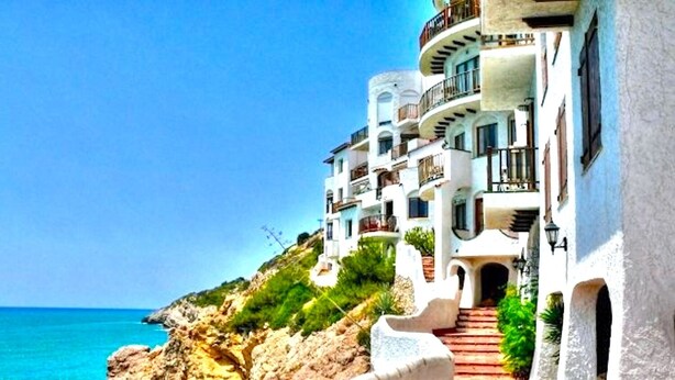 A coastal building with multiple balconies, a stone pathway leading to the entrance, and a view of the sea.
