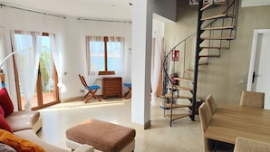 A spacious living area with a sofa, ottoman, and a dining table with chairs. A wooden staircase with a fire extinguisher is visible.