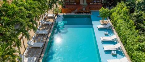 A rectangular swimming pool with lounge chairs and palm trees, surrounded by lush greenery and a building with a red-tiled roof.