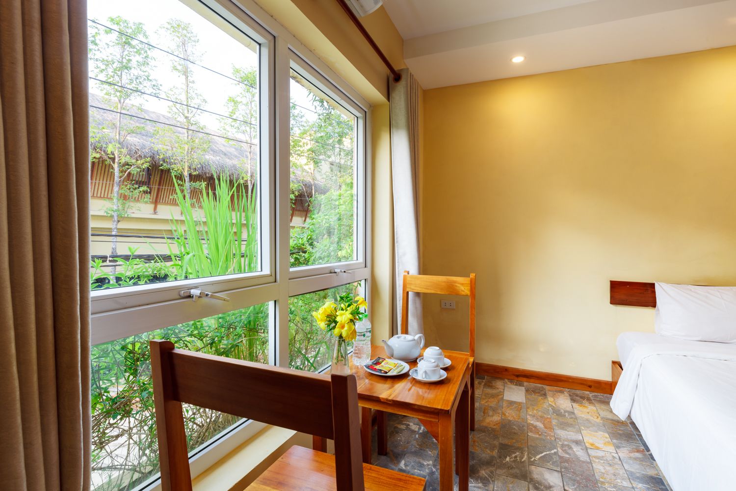 A room with a bed, a wooden chair, a small table with a vase of flowers, and a window with a view of greenery outside.