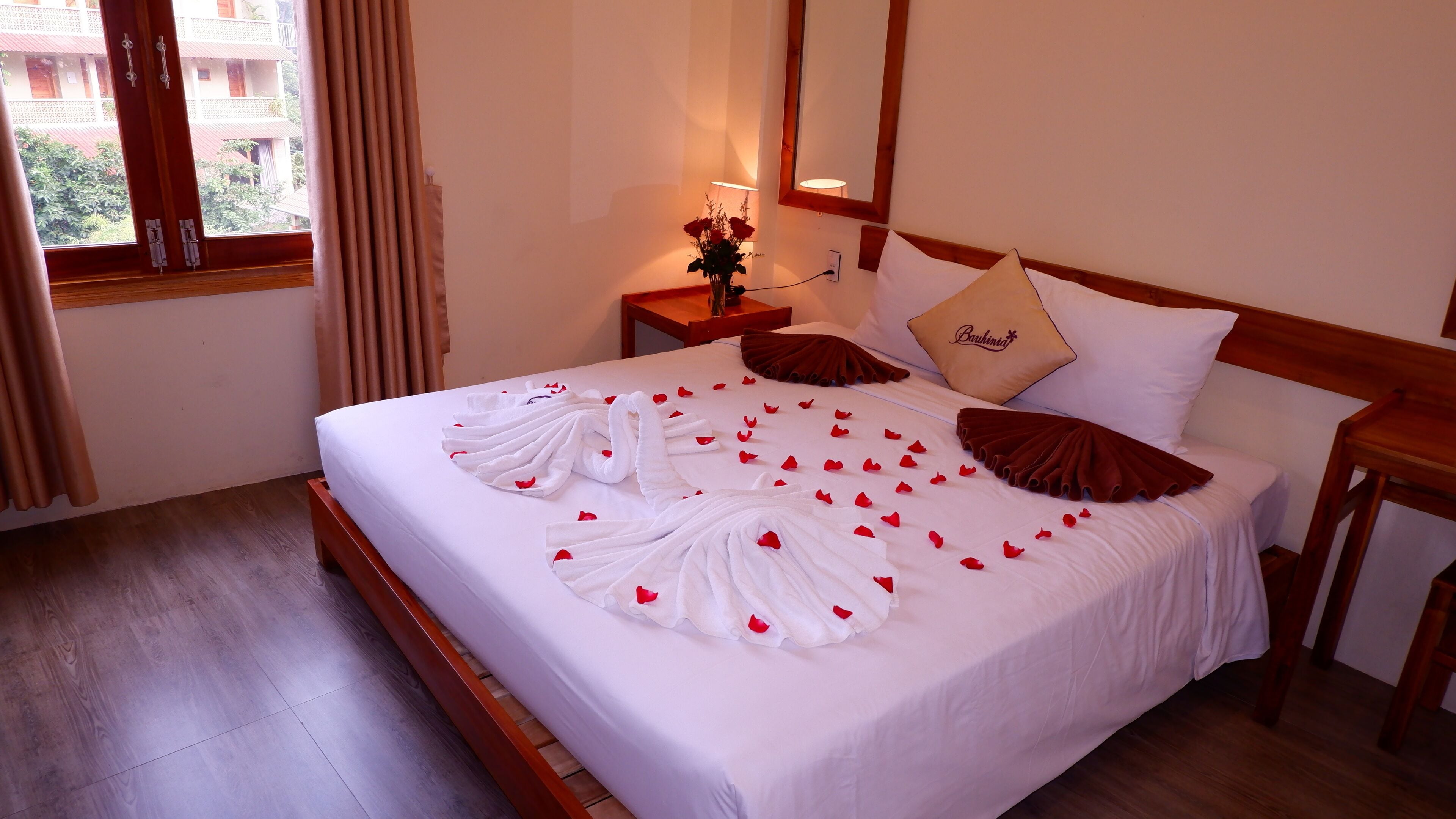 A hotel room with a neatly made bed, a nightstand with a lamp, a vase with flowers, and a window with curtains.