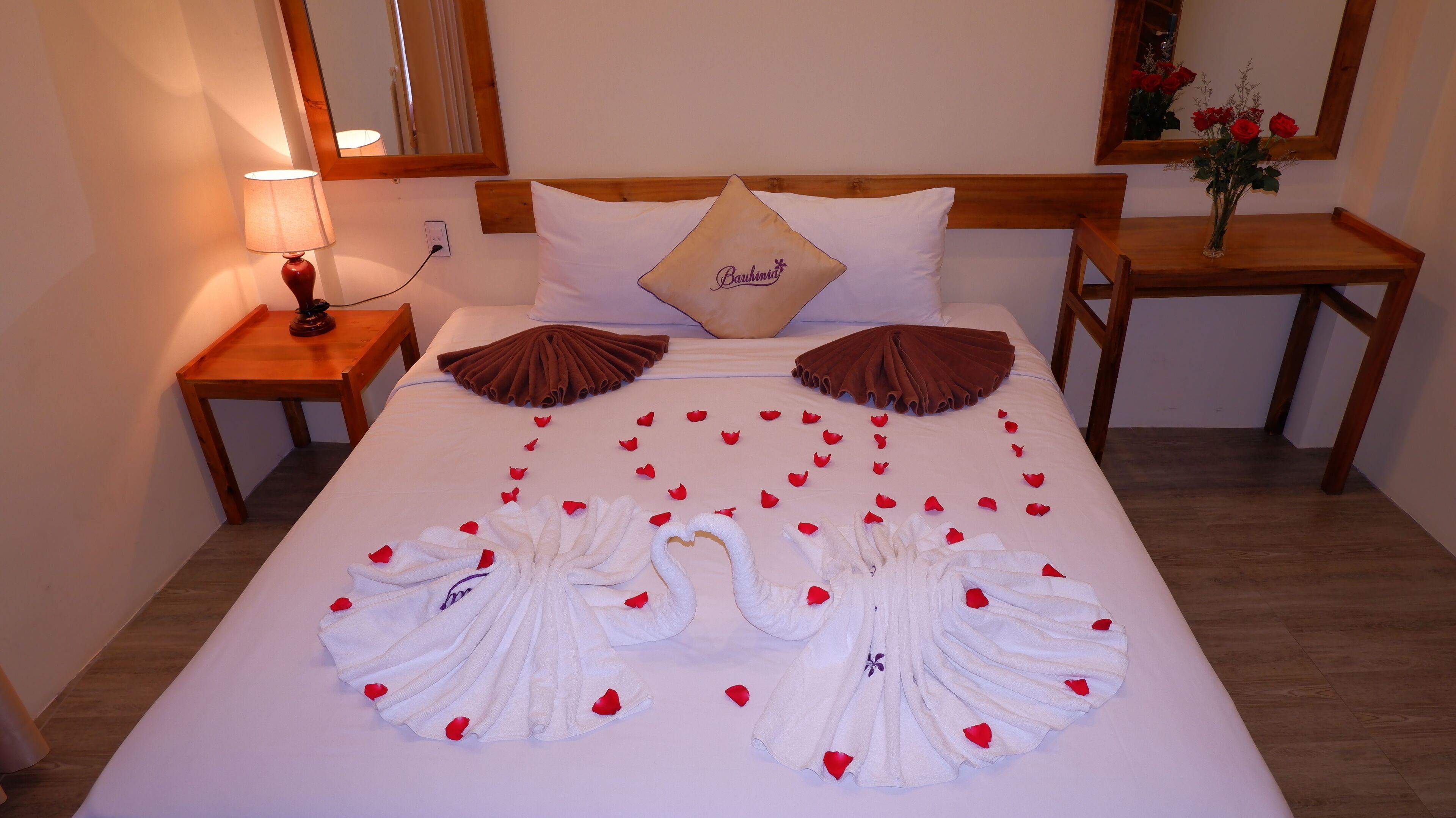 A neatly made bed with a heart-shaped towel arrangement and rose petals, a wooden headboard, a bedside table with a lamp, and a mirror on the wall.