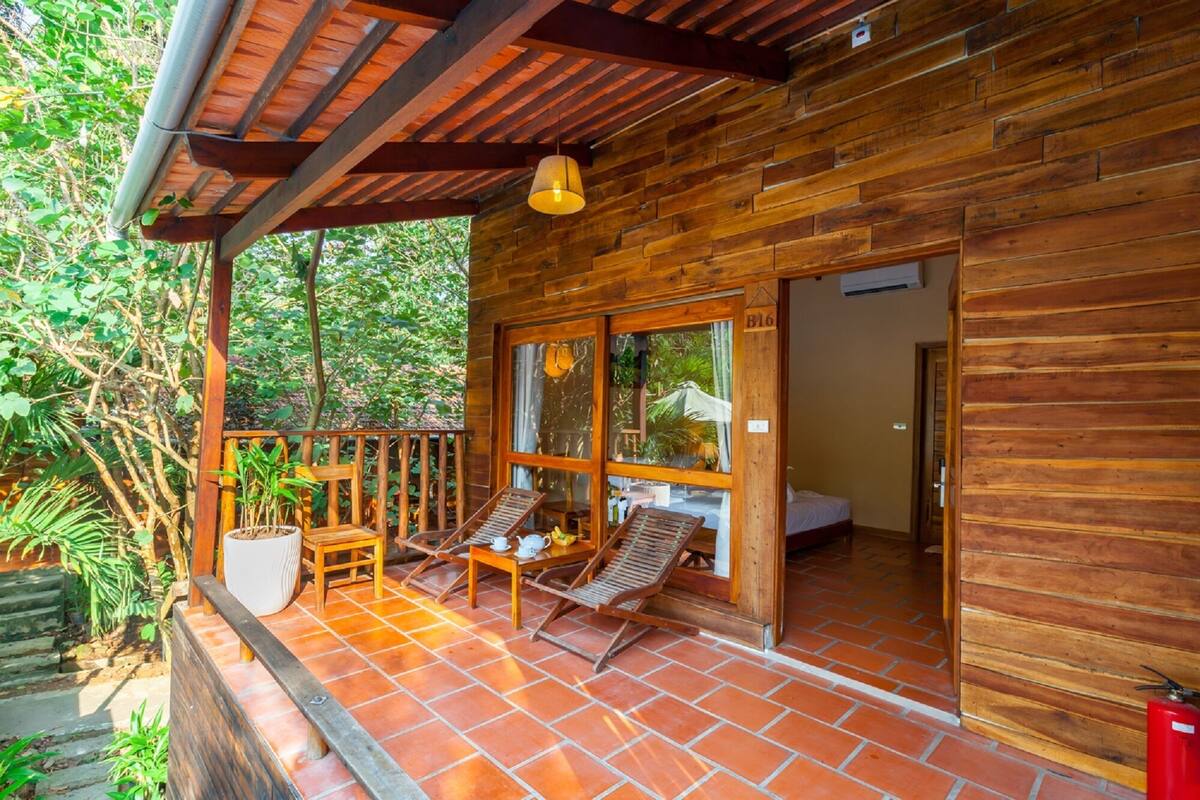 Una baita di legno con un patio piastrellato, un tavolino con sedie e una finestra che si affaccia sul verde.
