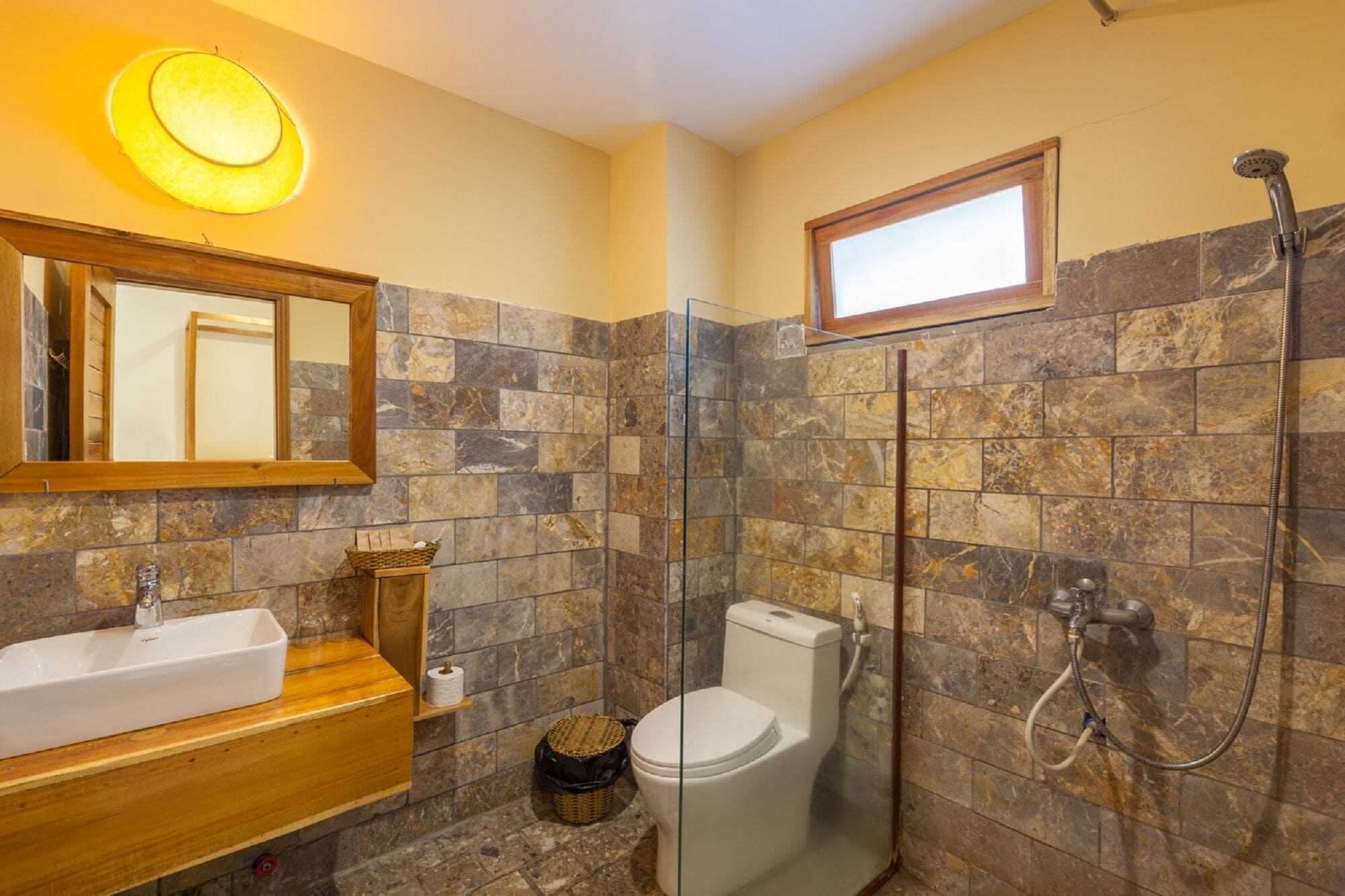 A bathroom with a stone tile wall, a wooden vanity, a white sink, a toilet, and a glass shower enclosure.