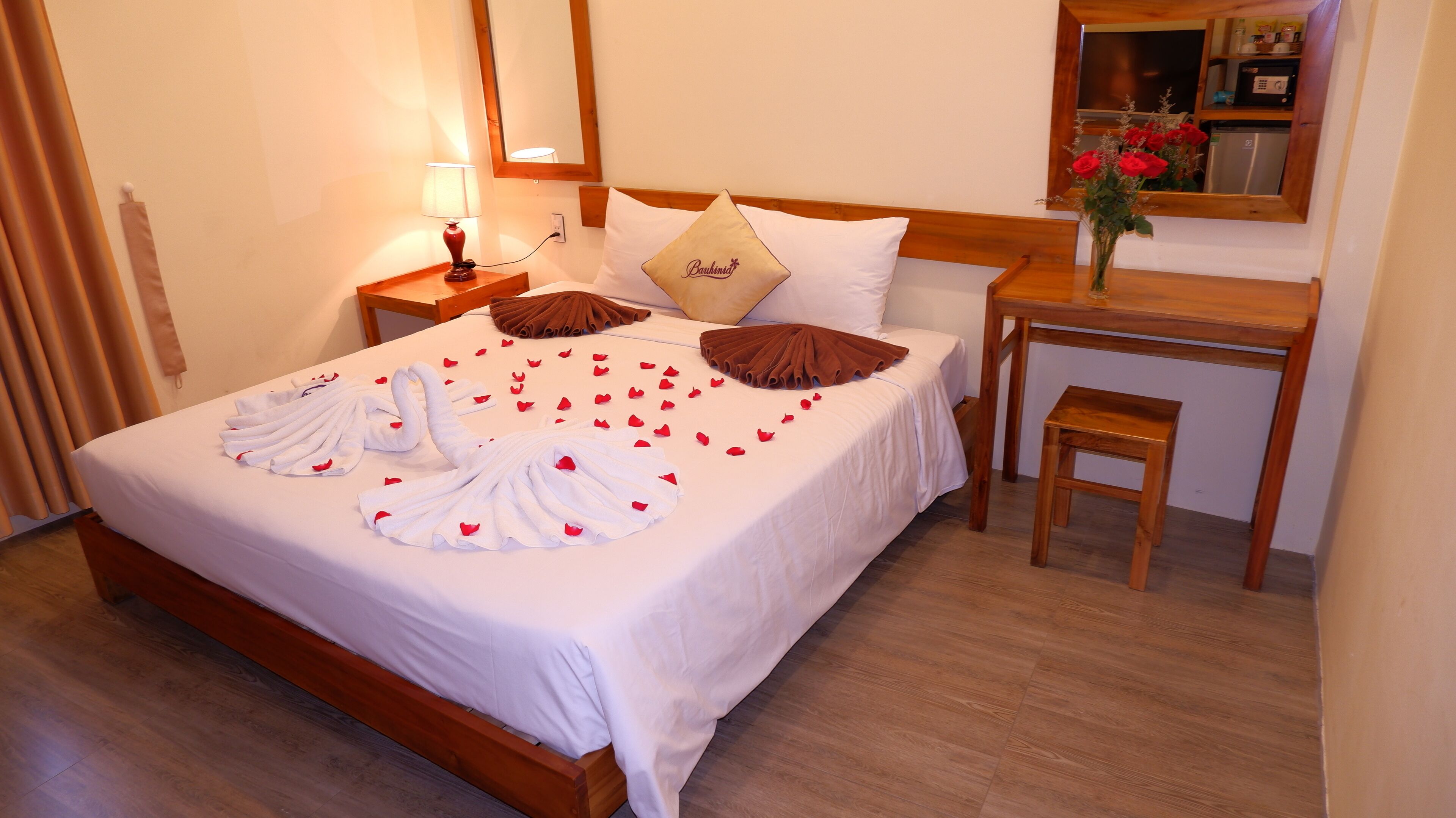 A hotel room with a neatly made bed, a bedside table with a lamp, a wooden nightstand, a small wooden stool, a mirror, and a vase with flowers.