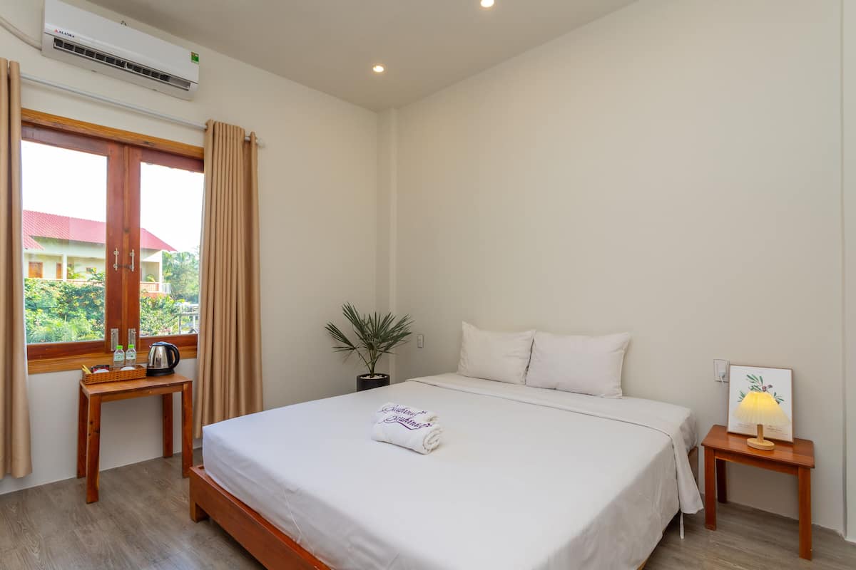 Una camera d'albergo ordinata con un letto, un comodino con una lampada, un tavolino con un bollitore e delle bottiglie, una pianta in vaso e una finestra con tende.