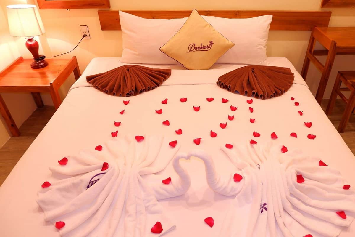 Una camera d'albergo con un letto ben rifatto, un cuscino ricamato con 'Bauhiniya' e petali di rosa disposti sul letto.
