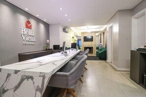 A hotel reception area with a marble counter, grey upholstered chairs, a flat-screen TV, and a wall-mounted sign reading 'Bella Vienna Pousada'. - Pousada Aconchego da Borges (Gramado)