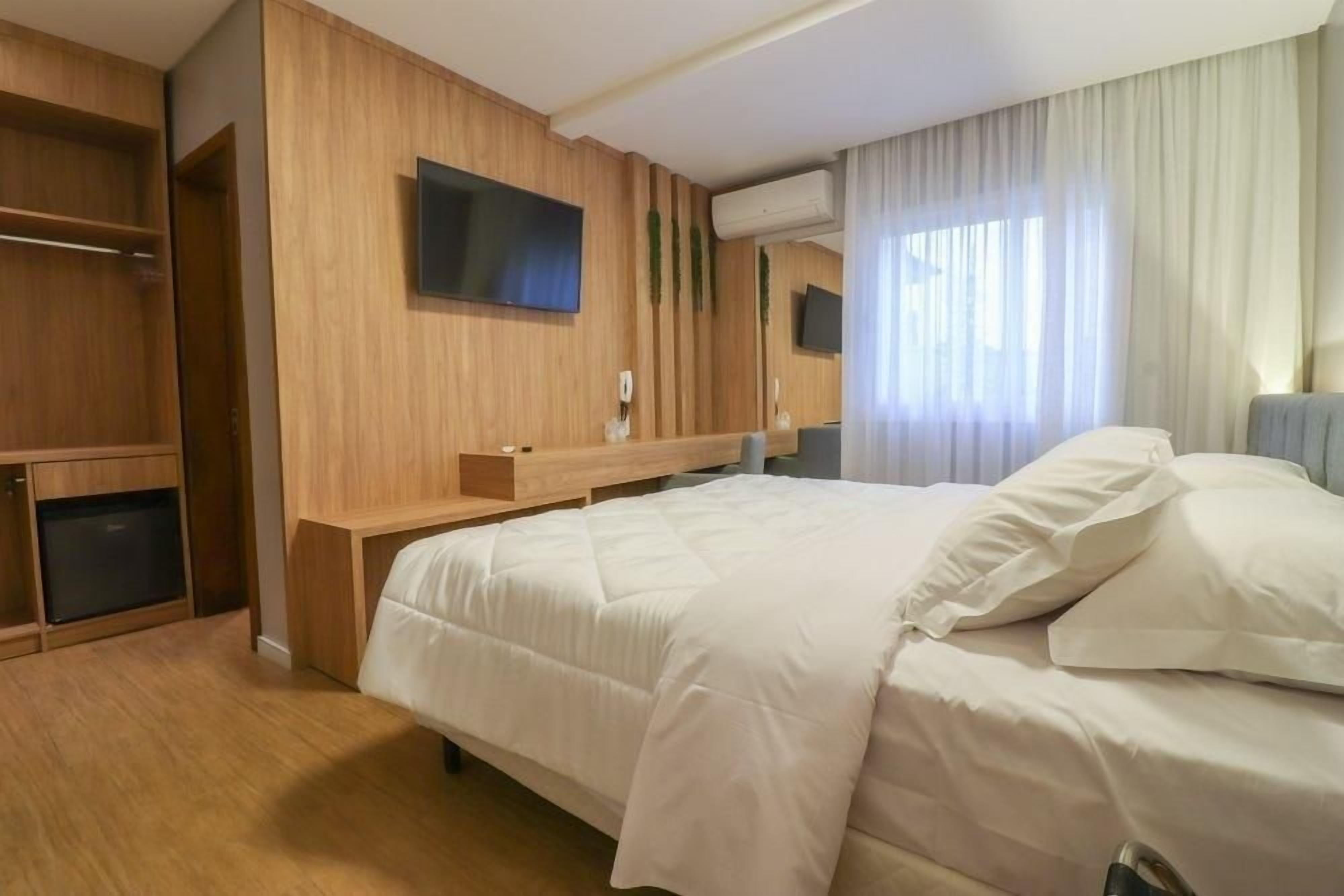 A hotel room with a bed, a TV mounted on the wall, a small desk, and a wardrobe.