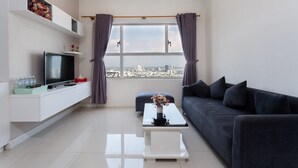 Un soggiorno moderno con una TV a schermo piatto, un tavolino bianco con un vaso di fiori, un divano scuro e vista sulla città attraverso ampie finestre.
