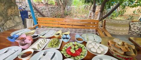 A wooden table set for a meal with various dishes, including olives, grapes, and bread, in an outdoor setting with trees and a stone wall in the background.