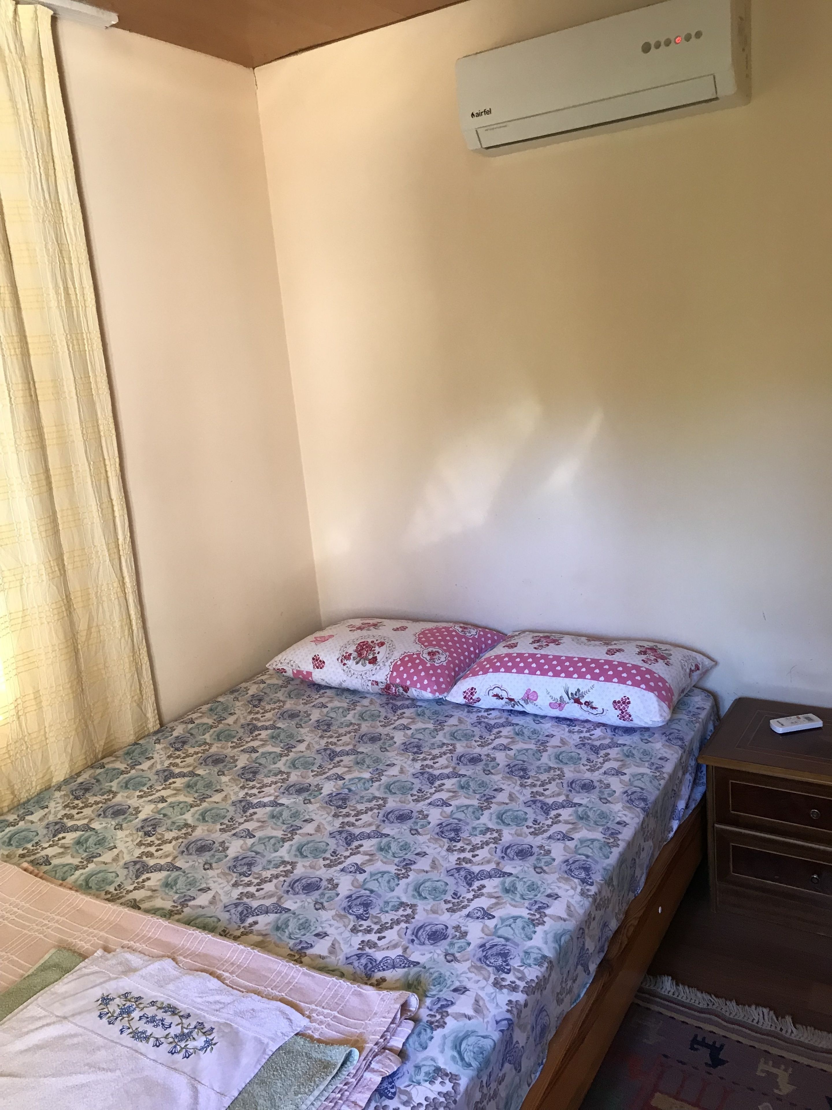 A small, single-bed room with a floral bedspread, a bedside table with a remote control, and an air conditioning unit on the wall.