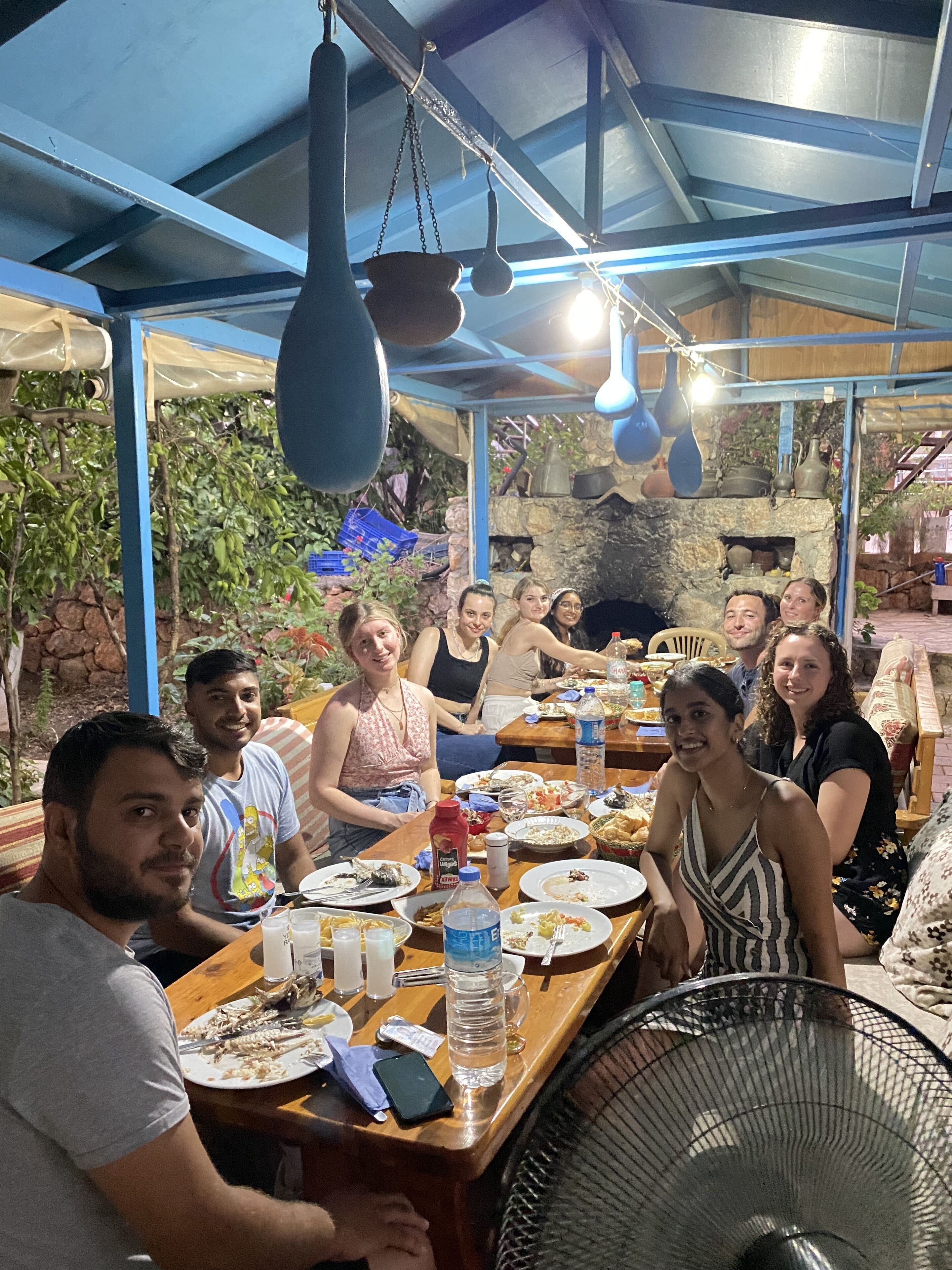Un groupe de personnes est assis à une table dans un restaurant en plein air, avec un mur de pierres en arrière-plan et des décorations suspendues.