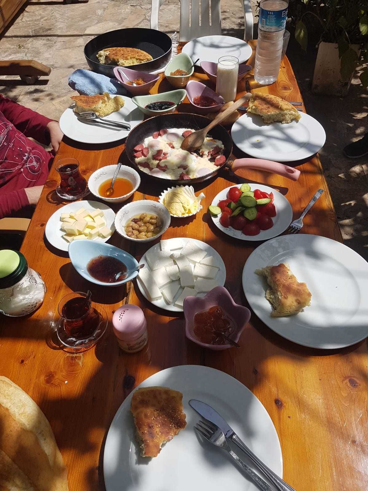 A wooden table set with various dishes including a pan with eggs and sausage, plates of cheese, olives, and a salad, along with drinks and utensils.