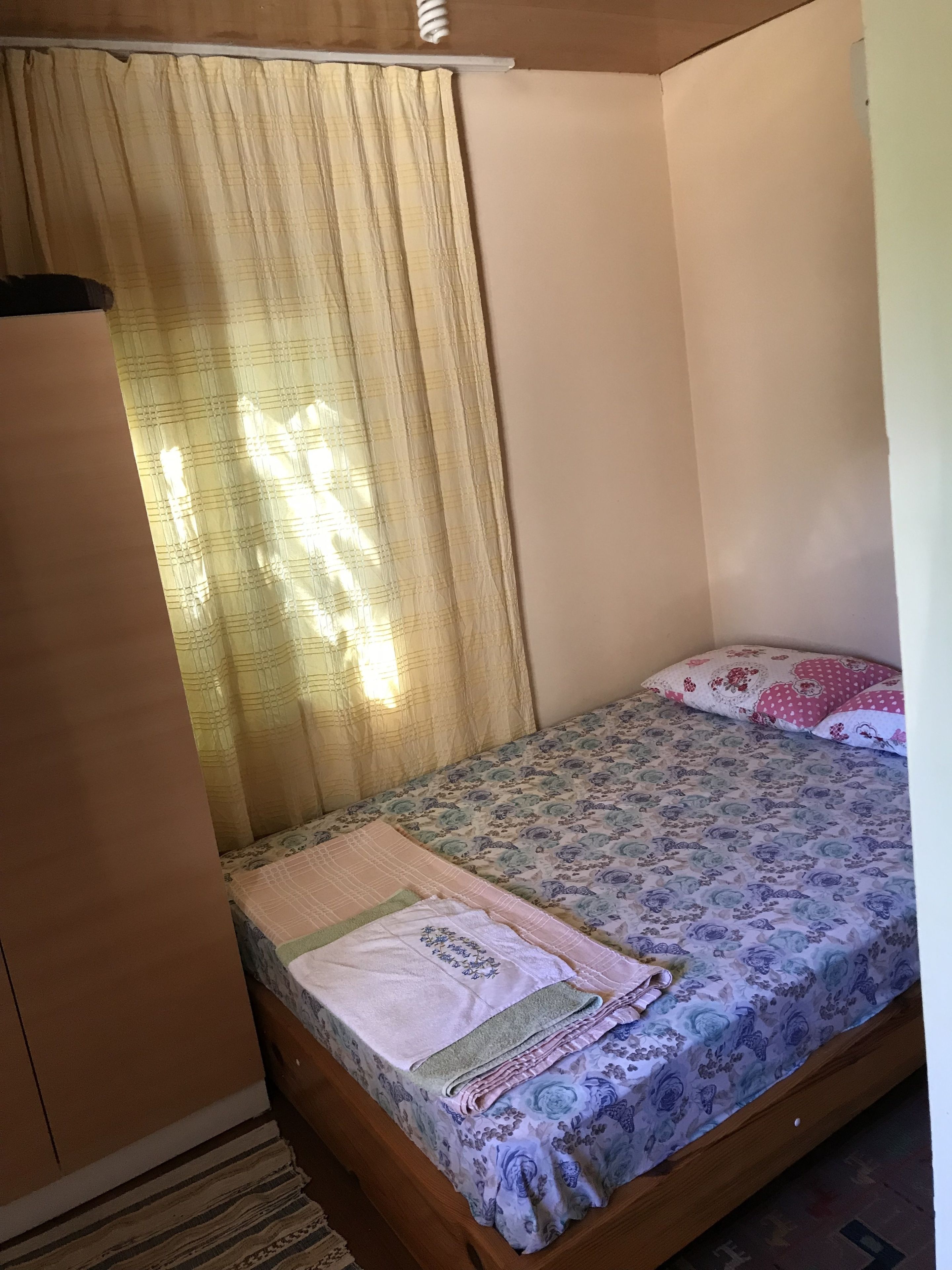 A small, single-bed room with a wooden headboard, a patterned bedspread, and a plain wall.