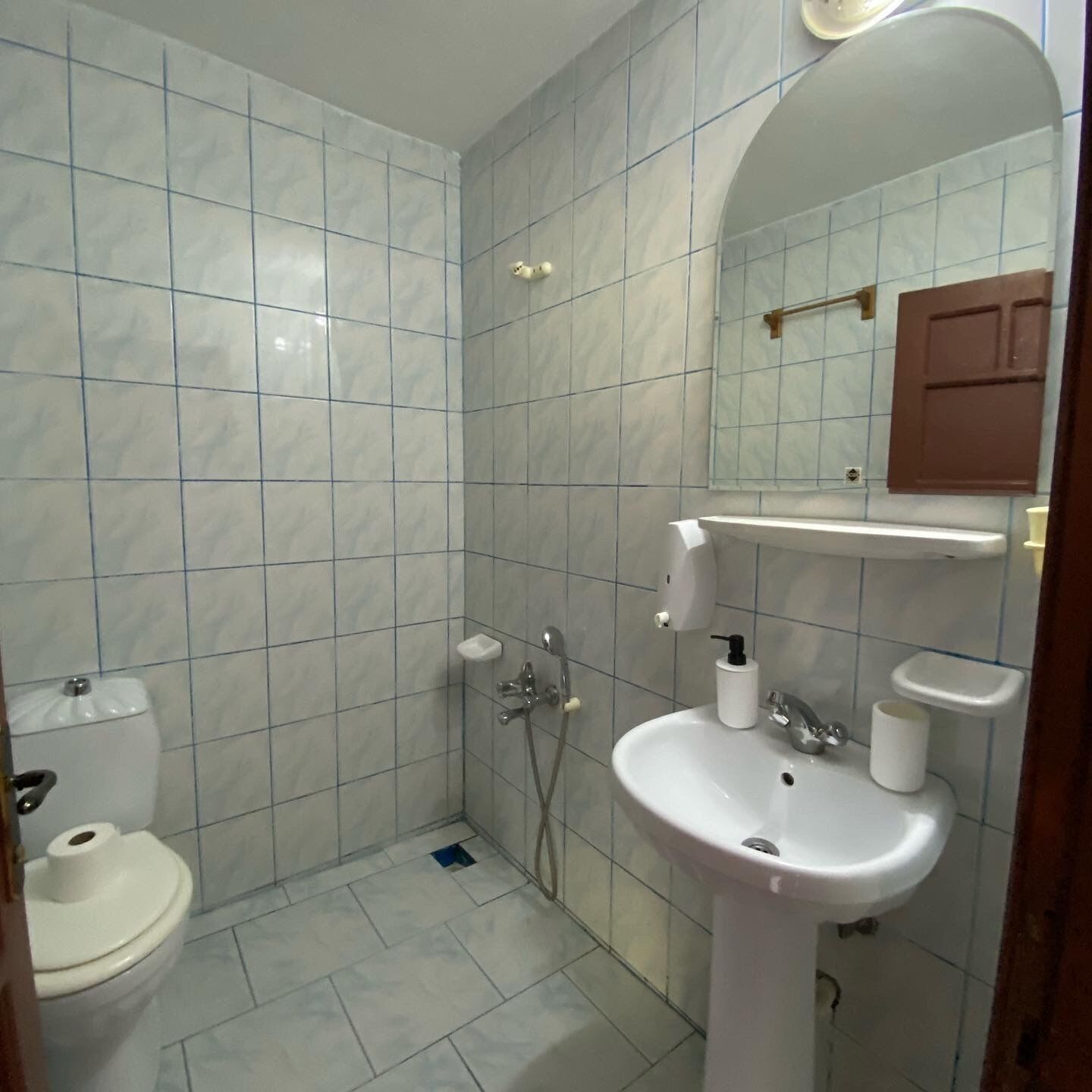 A bathroom with white tiles, a toilet, a sink with a mirror above, and a shower area.
