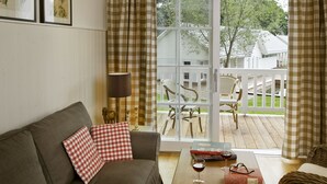 A living room with a sofa, a coffee table with books and glasses, a lamp, and a view of a deck with chairs and a house.