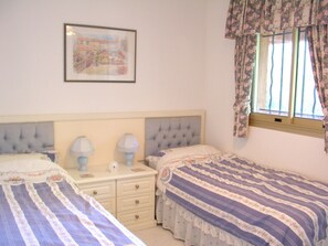 A bedroom with two beds, a nightstand, a framed picture on the wall, and a window with floral curtains.