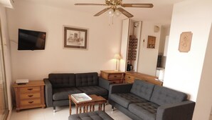 Een woonkamer met een grijze hoekbank, een houten salontafel, een aan de muur bevestigde televisie, een plafondventilator en een ingelijste foto.
