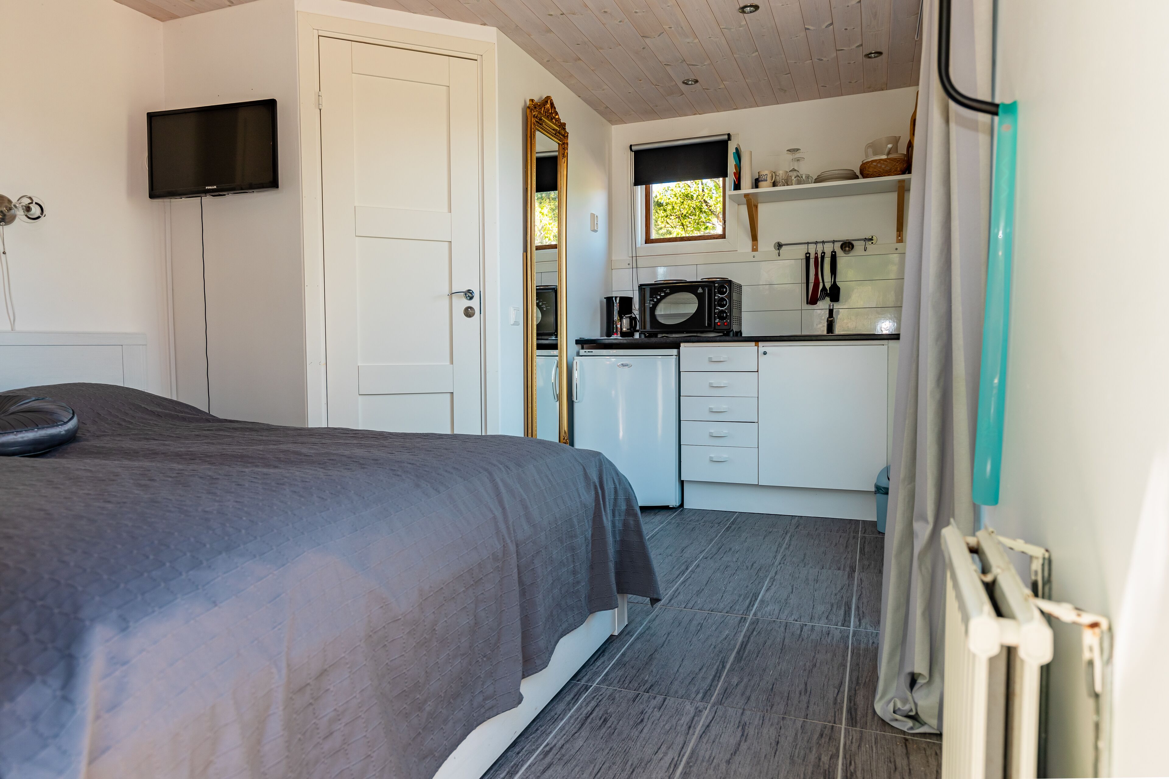 Een kleine kamer met een bed, een kitchenette, een televisie en een raam.