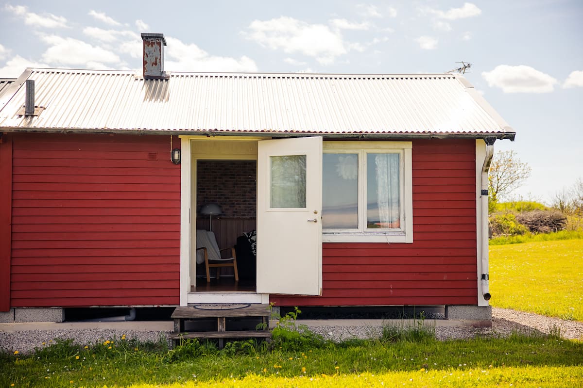 Uma casa térrea vermelha com uma porta e janela brancas, um pequeno alpendre e um muro de tijolos visível pela porta.