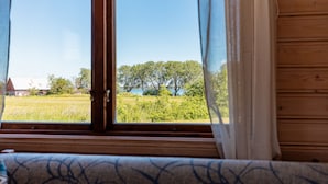 A window with curtains, a view of a green field, trees, and a building in the distance. - Fårösunds Semesterby (Fårösund)