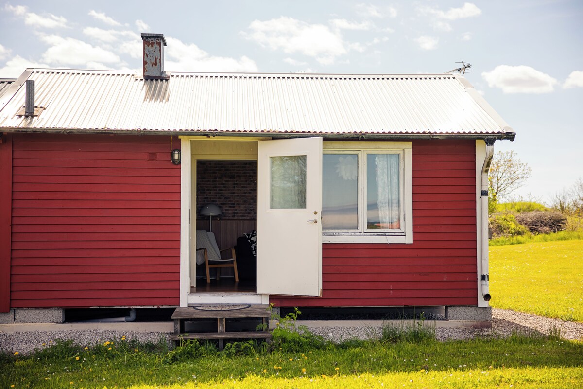 Una casa roja de un piso con puerta y ventana blancas, un porche pequeño y un muro de ladrillo visible a través de la puerta.