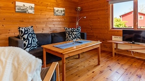 A cozy cabin interior with wooden walls, a sofa with patterned cushions, a wooden coffee table, a TV on a stand, and a window with a view of a red building. - Fårösunds Semesterby (Fårösund)