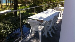 Una terraza con mesa y sillas blancas, rodeada por una barandilla de metal, con vistas a un jardín y una masa de agua.