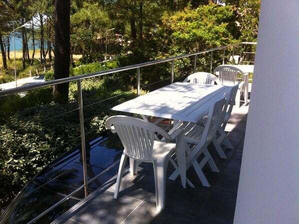 Una terraza con mesa y sillas blancas, rodeada por una barandilla de metal, con vistas a un jardín y una masa de agua.