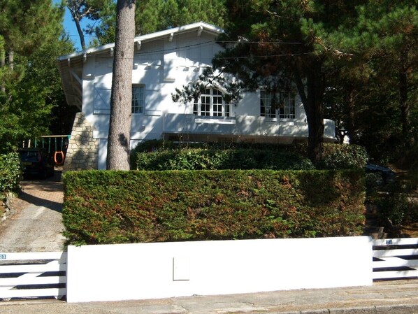 Une maison blanche aux fondations en pierre, entourée d’une haie et d’arbres.