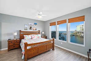 A bedroom with a wooden bed, two bedside tables, a ceiling fan, and large windows with a marina view.