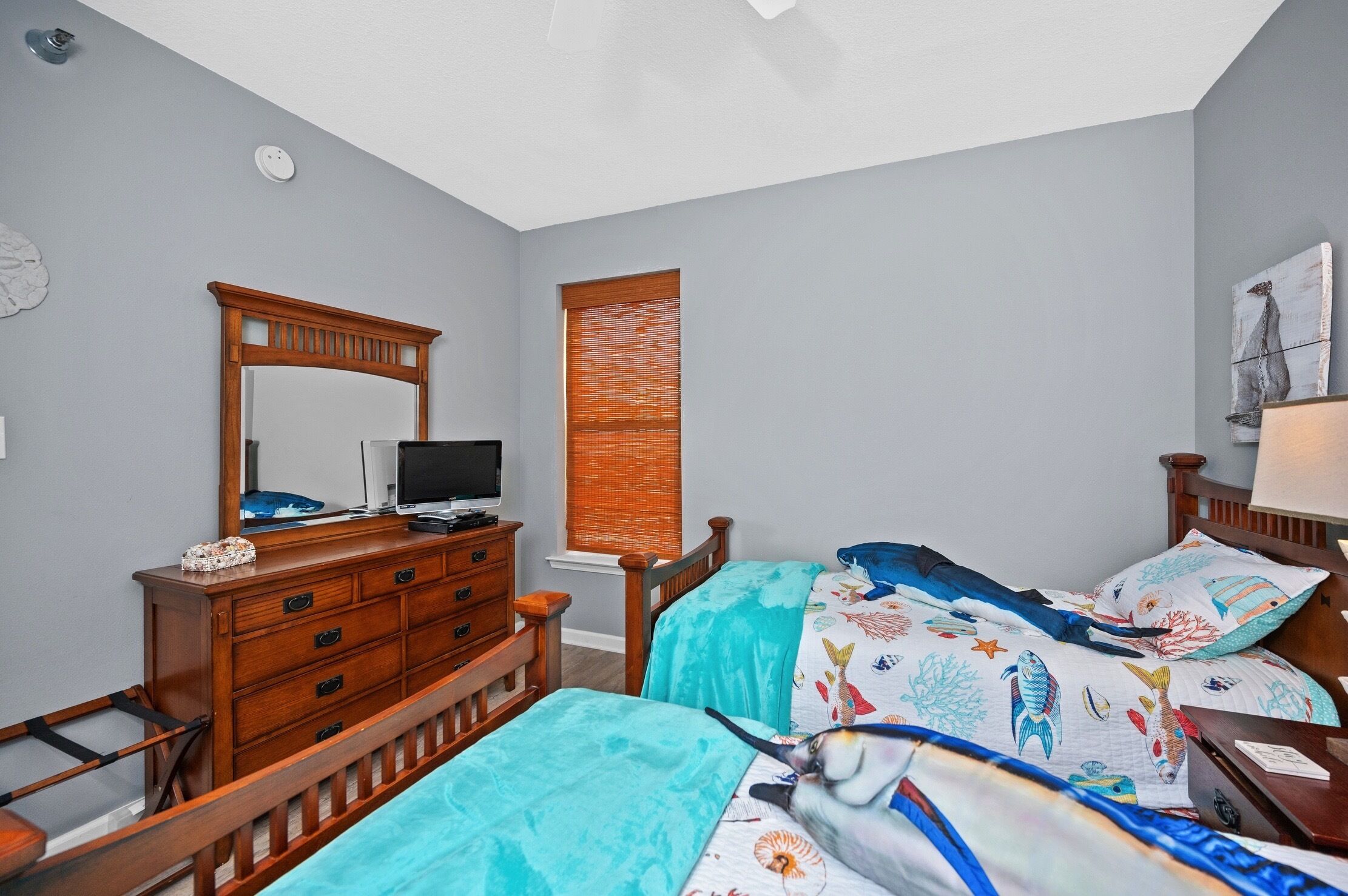 Un dormitorio con un aparador de madera, un televisor, una cama con colcha azul y blanca estampada, y una ventana con persianas.