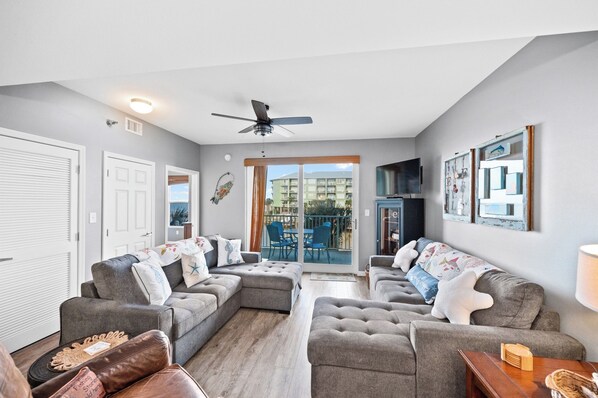 A living room with grey sofas, a ceiling fan, and a large window overlooking a balcony.