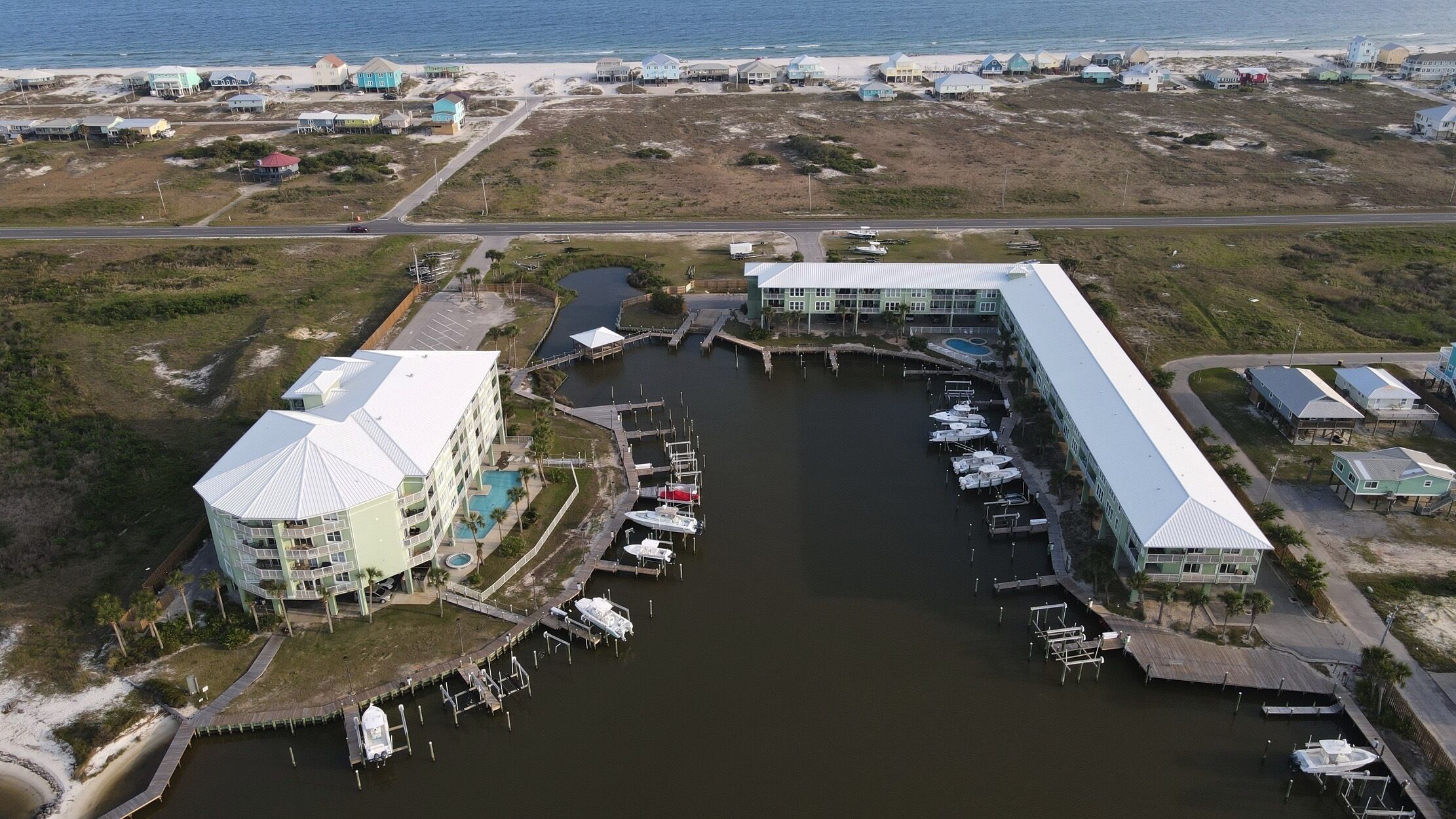 Una vista aerea di una marina con diverse imbarcazioni ormeggiate, un edificio grande con piscina e una spiaggia sullo sfondo.