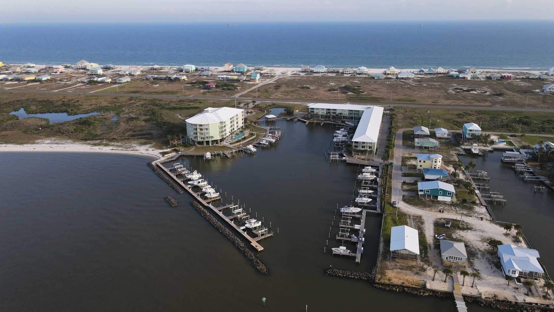 An aerial view of a coastal marina with multiple docks and boats, a large building with a flat roof, and a beach in the background.