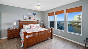 A bedroom with a wooden bed, two bedside tables, a ceiling fan, and large windows with a marina view.