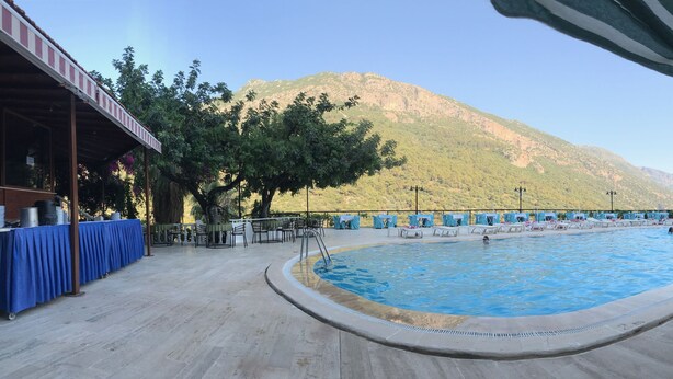 A swimming pool with a mountain backdrop, a covered seating area, and a table with stacked chairs.