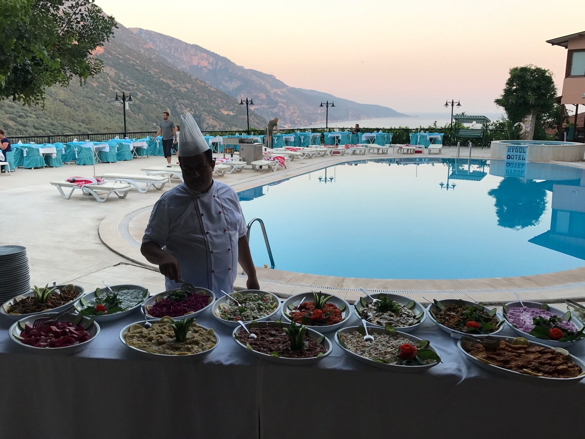 A chef serving food at a poolside buffet with a mountainous backdrop.