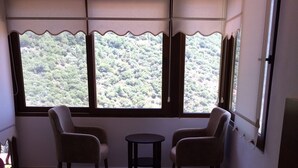 A hotel room with a bed, two chairs, a small table, and a view of a forest through large windows.