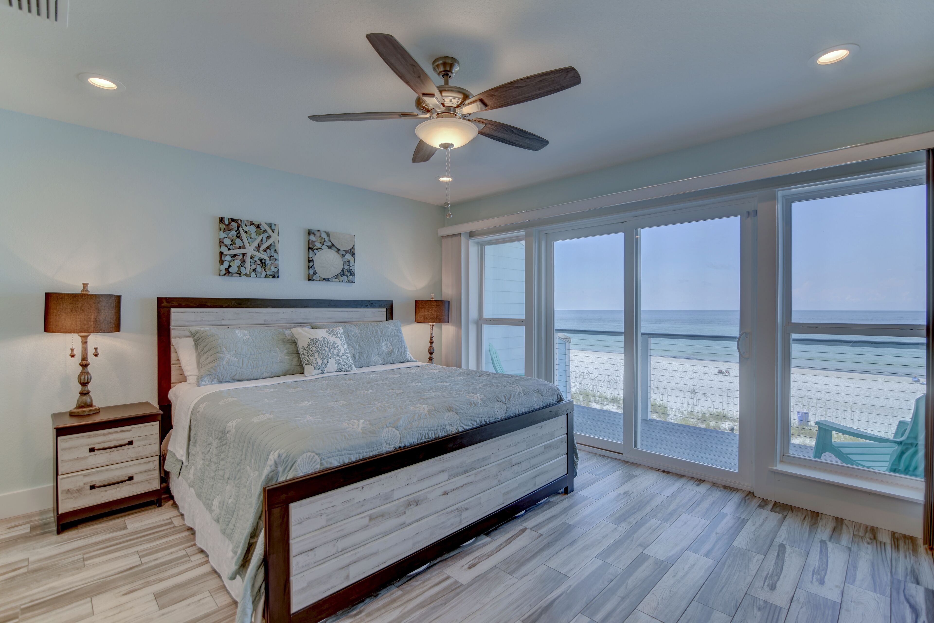 A bedroom with a large bed, wooden headboard, and a ceiling fan. It features a large window with a view of the beach.