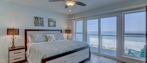 A bedroom with a large bed, wooden headboard, and a ceiling fan. It features a large window with a view of the beach.