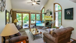 Ein geräumiges Wohnzimmer mit Deckenventilator, großen Fenstern, Kamin und Fernseher.
