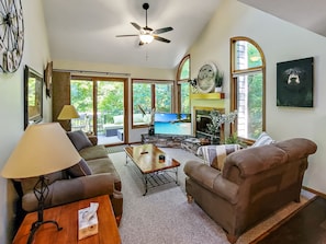 A spacious living room with a ceiling fan, large windows, a fireplace, and a television.