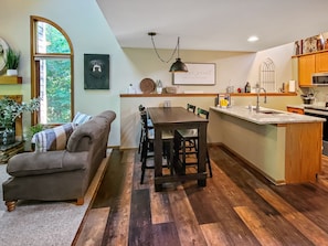 A cozy living room with a sofa and a dining table, a kitchen with wooden cabinets and a marble countertop, and a large window with a view of greenery.